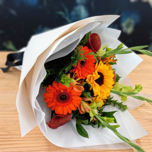 A wrapped bouquet of flowers featuring bold warm colors, including orange and yellow, with greenery.