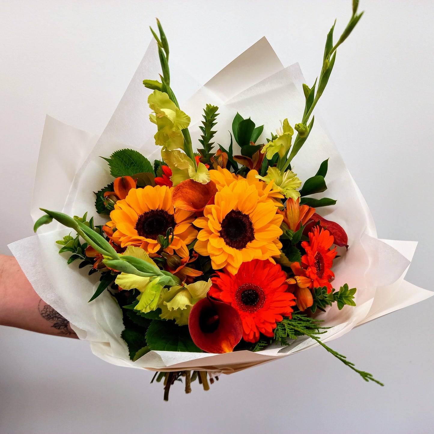 A bouquet of flowers with sunflowers, gerbera daisies, and other foliage wrapped in white paper.