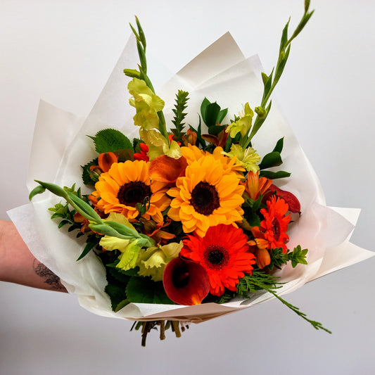 A bouquet of flowers with sunflowers, gerbera daisies, and other foliage wrapped in white paper.